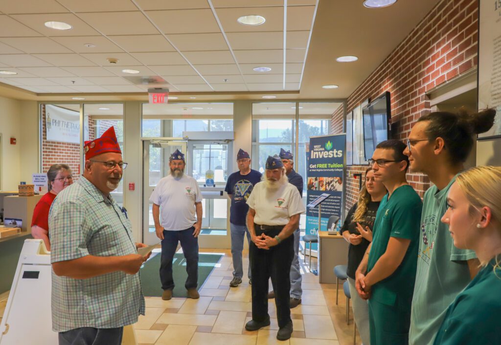 A group of individuals, including some in medical scrubs and one wearing a red hat, are standing in a well-lit room with beige tiled floors and brick walls. Posters are visible. A person stands near a sign.
