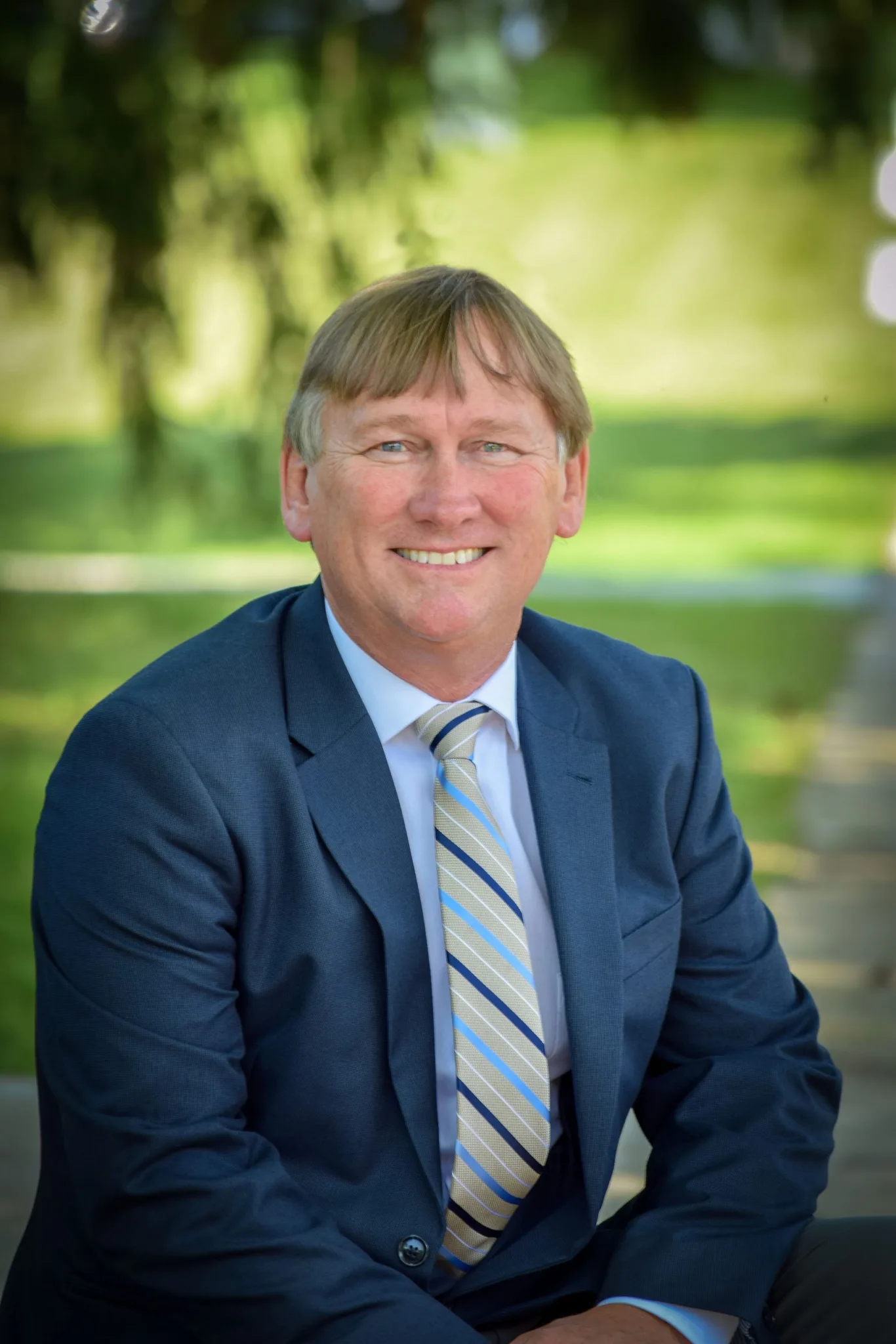 Portrait of a man wearing a suit sitting down with an outdoor scene behind him.