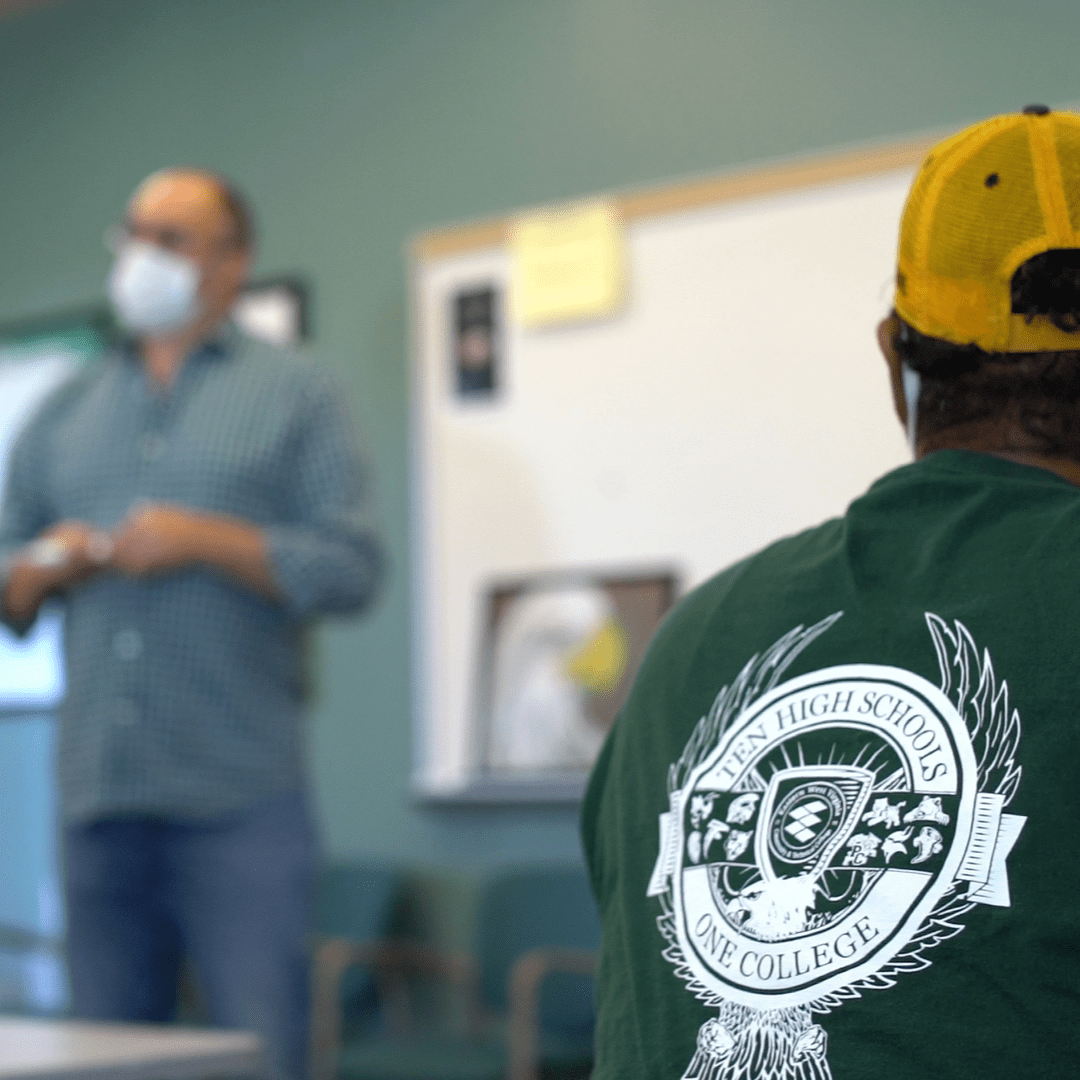 Photo of a man with a yellow hat, wearing a green shirt, watching a man standing.