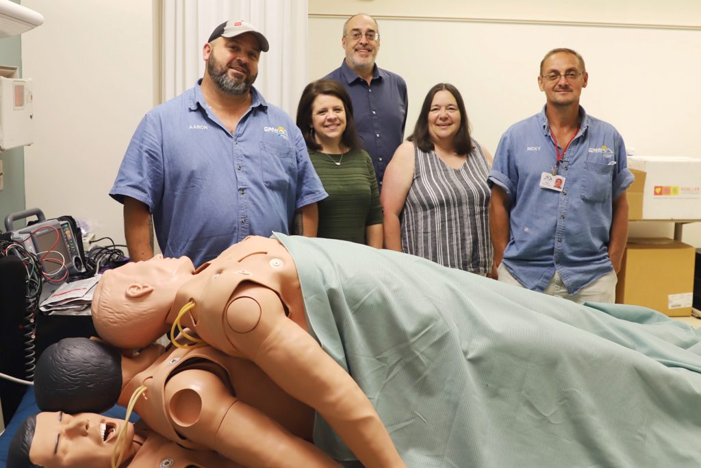 A group of people stand behind three nursing skills training mannequins.