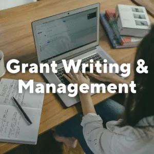 Photo of woman typing on a laptop with open journal by her. Text overlay, "Grant Writing & Management."