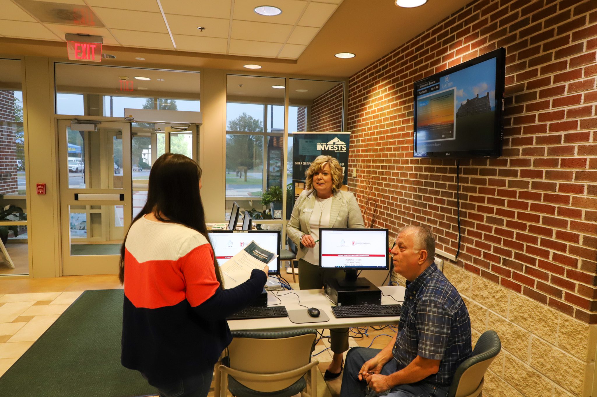 Photo of a woman speaking with a student and an older man sitting down at a computer.