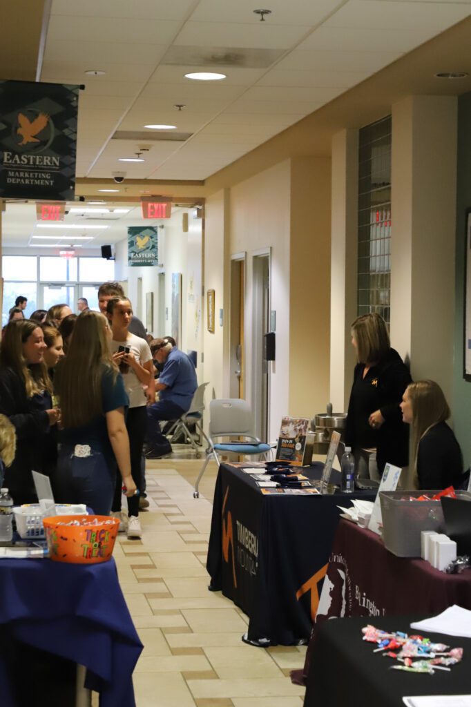 Photo of a group of people in a hallway. Tables set up throughout from different organizations.