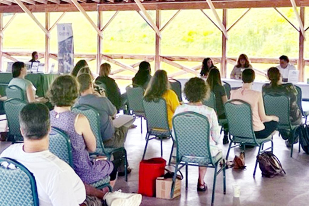 A group of people sitting on chairs listens to a group of speakers sitting at a table in front of the group.