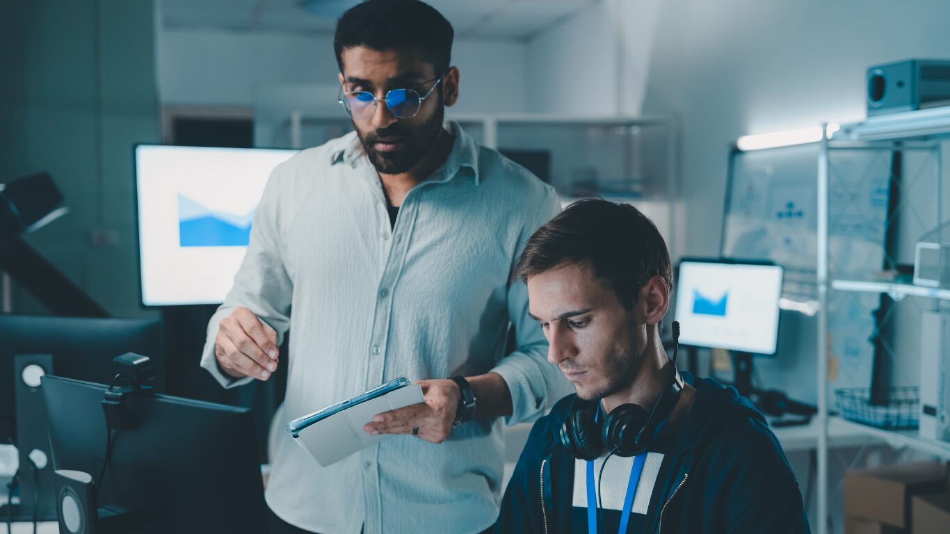Photo of two men in a room with computers. One man is holding a tablet and the other has headphones around his neck, looking down toward a computer.