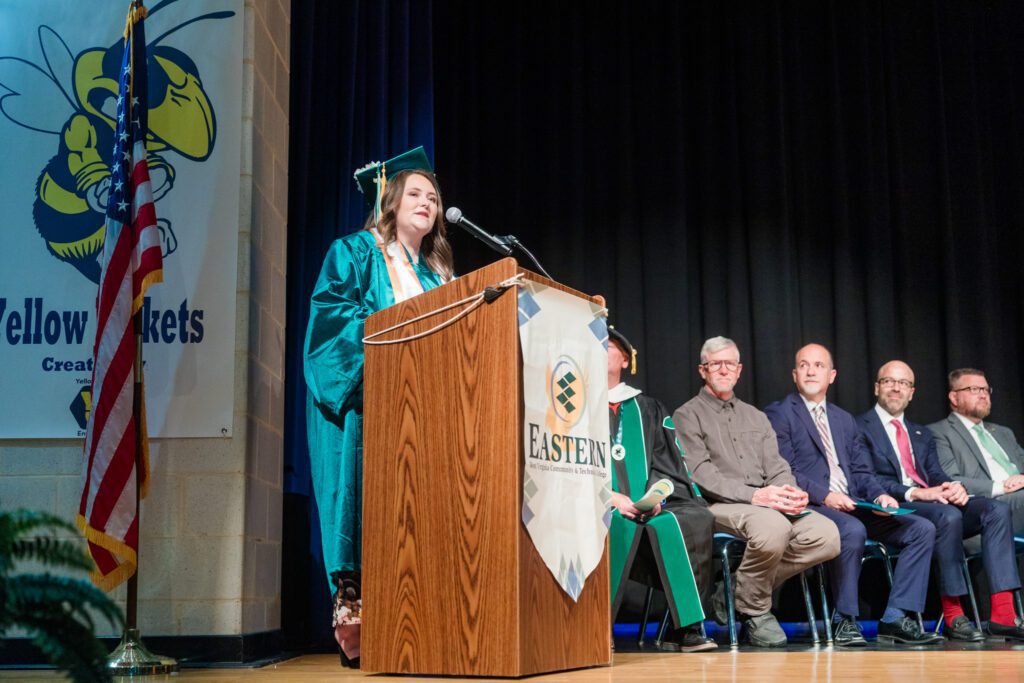 Photo of Miranda Simon at podium on a state with others sitting behind her.