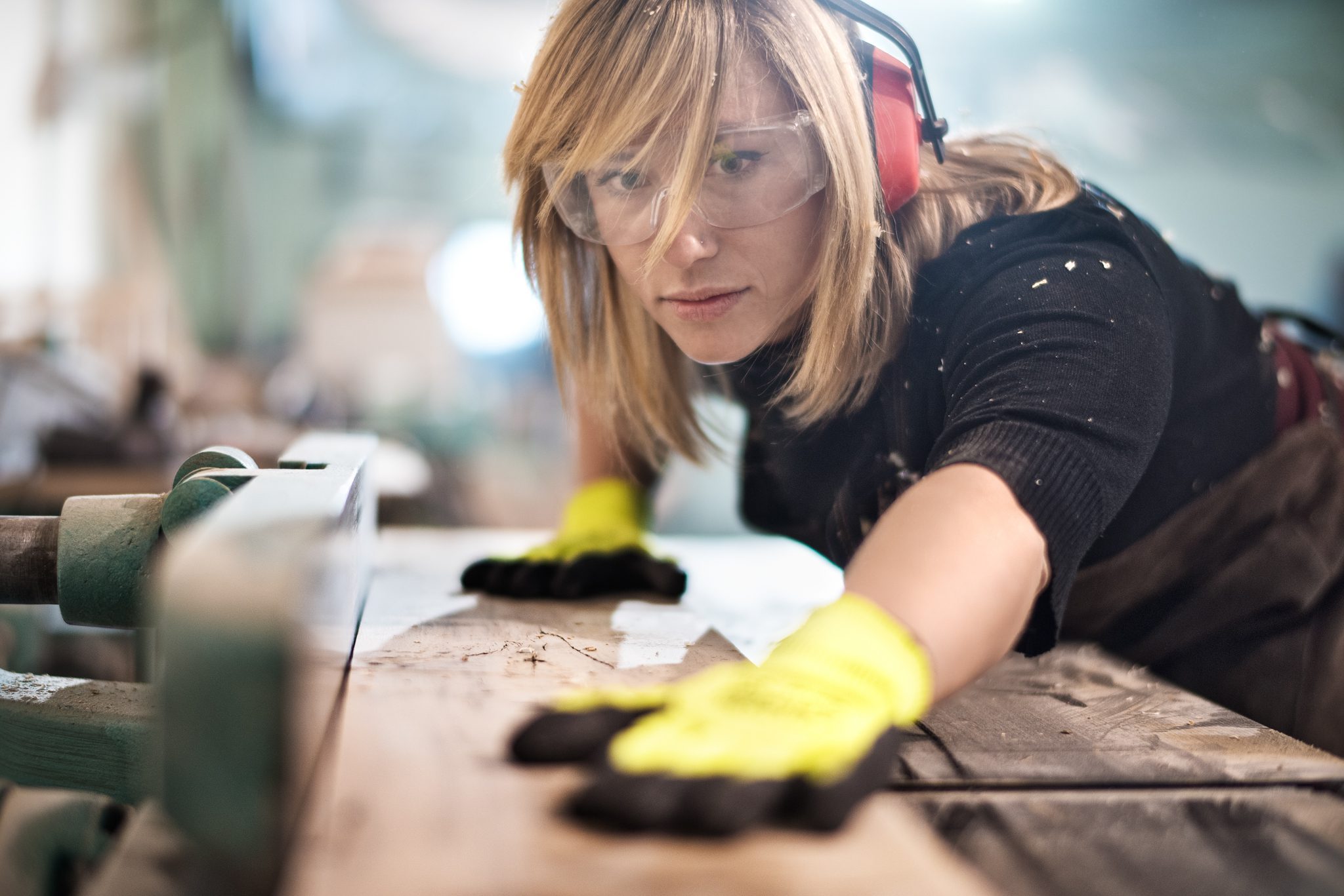 Female craftperson with protective equipment working with planks in workshop. She is making furniture parts.
