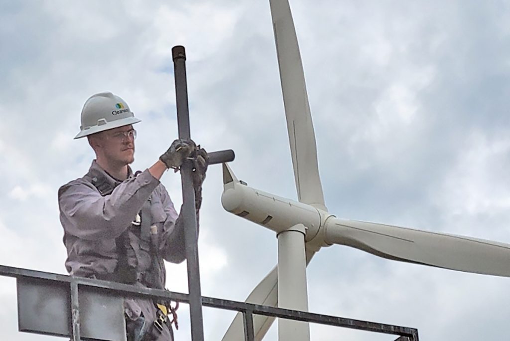 A man wearing a hard hat works on a platform. A wind turbine is behind him.